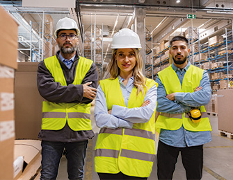 Tres trabajadores en un almacén, usando cascos y chalecos amarillos, con un fondo de estanterías y cajas.