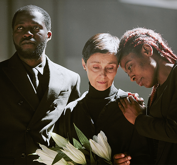 Tres personas vestidas de negro están en un entorno iluminado. Una mujer sostiene un ramo de lirios, mientras otra la abraza. Un hombre observa con expresión seria.