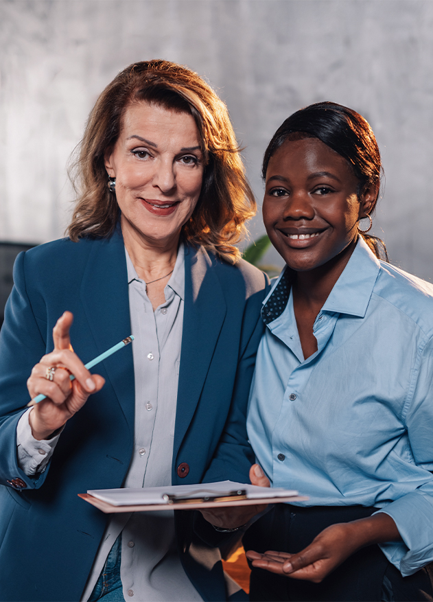 Dos mujeres sonrientes en un entorno de oficina. Una lleva un blazer azul y sostiene un lápiz, mientras la otra viste una camisa azul claro. Ambas muestran un cuaderno.