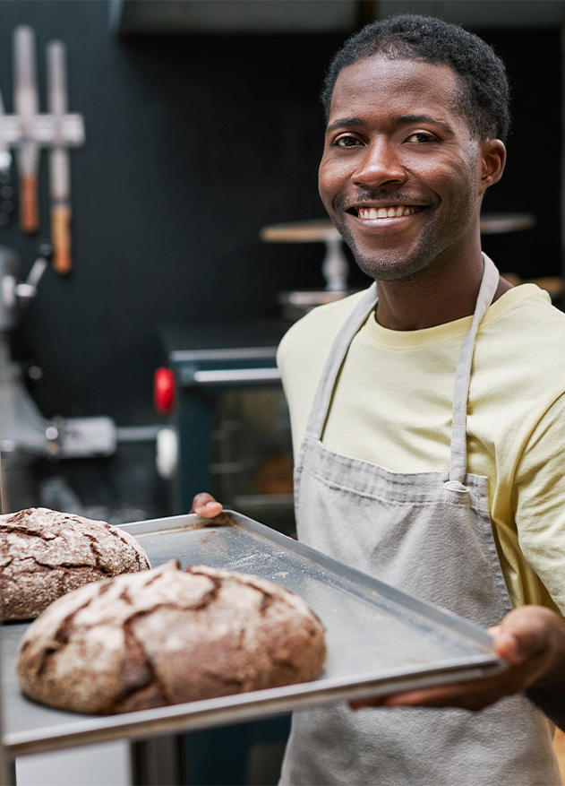 Hombre sonriente sostiene una bandeja con dos panes recién horneados en una panadería. Fondo oscuro y utensilios de cocina visibles.
