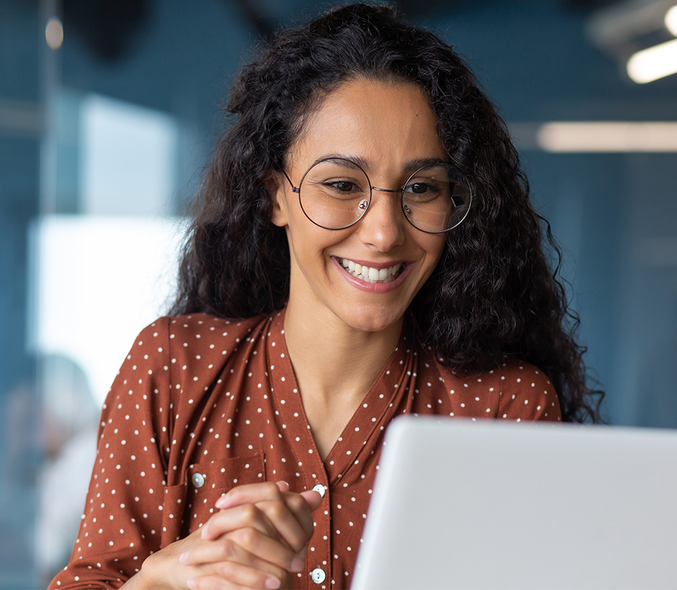 Mujer con cabello rizado y gafas, sonríe mientras observa una computadora portátil. Lleva una blusa marrón con puntos blancos. Fondo de oficina con tonos azules y luz natural.
